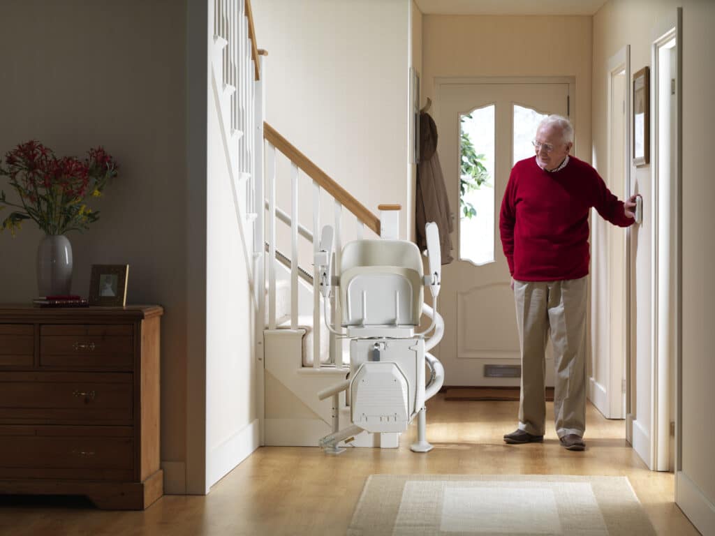 Man using a wall mounted remote control to call a folded cream stairlift in a bright hallway.
