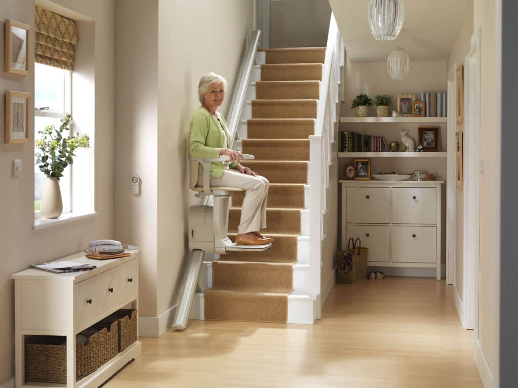 The woman sits on the stairlift as it travels up the first few steps of the staircase. She is looking towards the camera with a smile while resting her hands on the armrests.