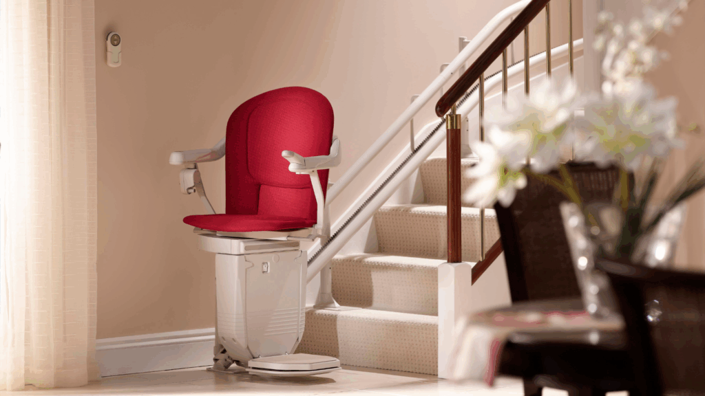A white curved stairlift with a red seat parked at the bottom of a carpeted staircase. A small, circular remote control unit is mounted on the wall nearby.