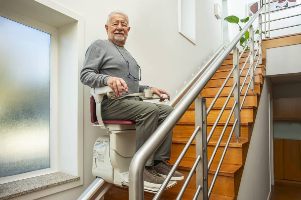 A side-view profile of an older man with a grey beard and sweater descending the wooden stairs on the white and red stairlift, looking toward the camera.