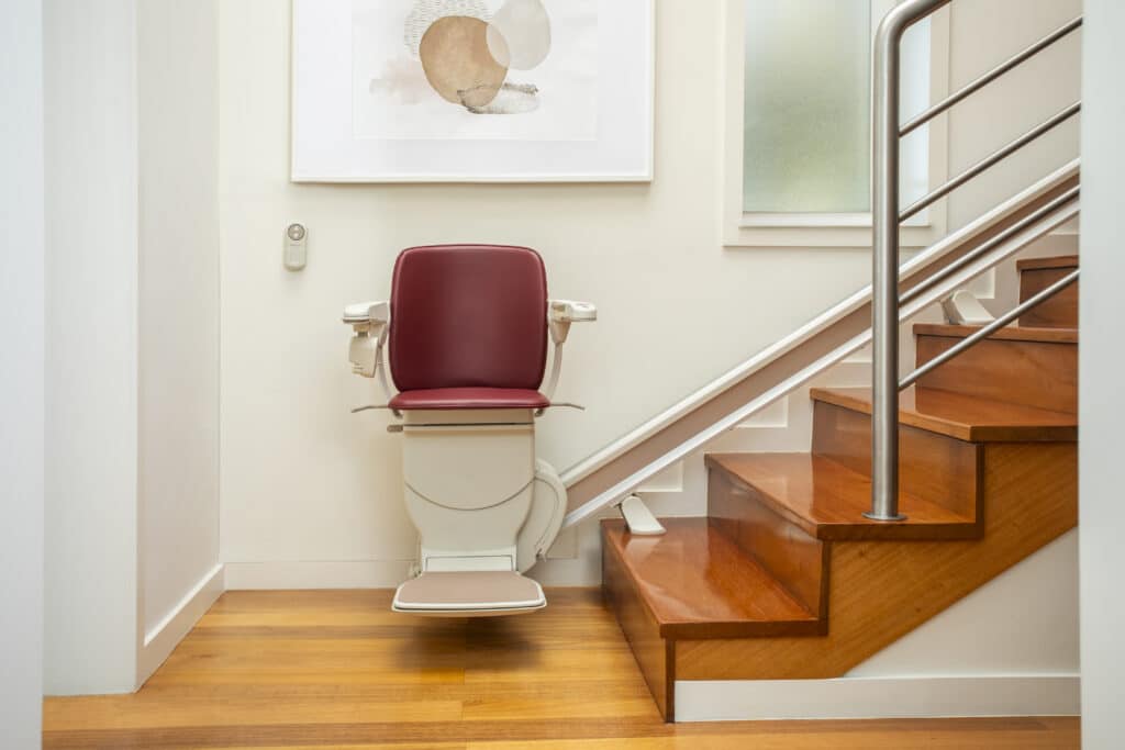 A clear front view of a white stairlift with a red seat parked at the bottom of a bright, modern wooden staircase. A wall-mounted remote control is visible on the left.