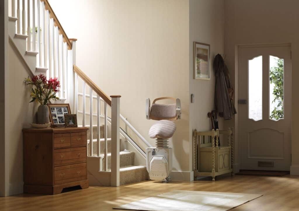 A white curved stairlift with a folded footrest at the base of carpeted stairs, with a wooden chest of drawers nearby.
