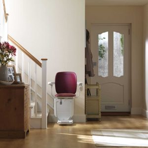 Red seated curved stairlift positioned at the base of a staircase next to a wooden chest of drawers.