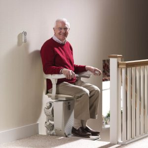 Older man sitting on a cream upholstered stairlift at the top of a carpeted staircase next to a banister.