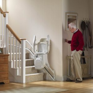 Man in a hallway using a remote control for a stairlift parked further up the steps.