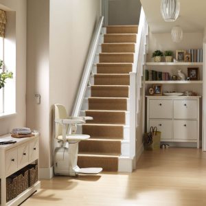 The cream coloured stairlift is unfolded at the bottom of the straight staircase with the seat and footrest in the lowered position. The hallway features light wood flooring and a white cupboard with baskets underneath.