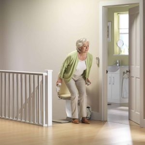 An older woman in a green cardigan stands up from a cream coloured stairlift at the top of a staircase. She is holding onto the armrests for support while an open door in the background shows a brightly lit bathroom.