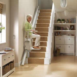The woman sits on the stairlift as it travels up the first few steps of the staircase. She is looking towards the camera with a smile while resting her hands on the armrests.