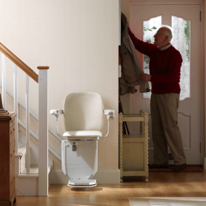 A cream-coloured stairlift parked at the bottom of a staircase in a well-lit hallway. The man from the previous image is standing nearby, hanging his coat on a rack by the door.