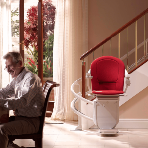 A white curved stairlift with a red seat parked at the bottom of a staircase. In the foreground, a man sits at a dining table reading a newspaper, with a bright window behind him.