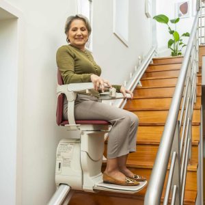 A side-view profile of the woman using the stairlift to descend the wooden stairs. Technical information and weight capacity labels are visible on the side of the white carriage base.