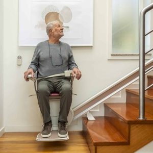 The man sits on the stairlift at the bottom of the staircase, looking upward. A modern stainless steel handrail runs alongside the wooden steps.