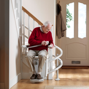 An elderly man wearing a red sweater sits on a white curved stairlift in a hallway, pulling the seatbelt across to secure himself before use.
