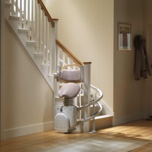 A white curved stairlift featuring a patterned saddle seat and folded footrest, parked at the base of a staircase in a bright hallway.