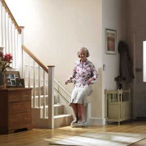 An older woman with a smile, wearing a floral blouse and beige skirt, seated on a white stairlift at the bottom of a flight of stairs.