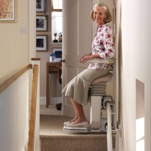An older woman seated on a stairlift at the top landing of a staircase, looking towards an open doorway.